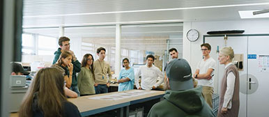 Eine Gruppe von Studierenden ist an einem Tisch versammelt und hört sich eine Präsentation an.