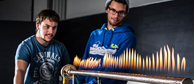 Zwei Studierende bei einem Schwingungsexperiment an einem Rohr mit vielen Gasflammen