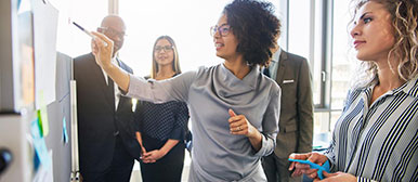 Eine Frau erklärt etwas an einem Whiteboard anderen umstehenden Personen