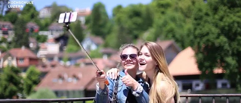 zwei junge Frauen machen ein Selfie mit dem Selfiestick, im Hintergrund diverse Gebäude und Bäume