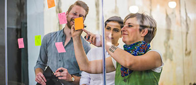 Eine Frau klebt Post its auf eine Glaswand, zwei weitere Personen sehen zu