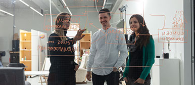 Drei Studierende stehen im Büro hinter einer Glasfront, auf der ein Mindmap aufgezeichnet ist und von ihnen analysiert wird