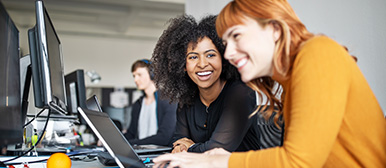 Eine Studentin in gelbem Pulolover und eine in schwarzem Pullover arbeiten lächelnd vor ihren Bildschirmen
