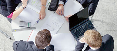 Eine Gruppe Studierender lernen an einem runden Tisch, der mit Blättern und Laptops bedeckt ist, aus Vogelperspektive fotografiert