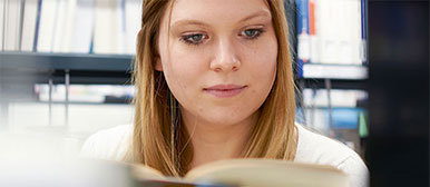 Eine Studentin von nah aufgenommen, liest konzentriert in einem Buch und im Hintergrund ist ein Bücherregal voller Bücher