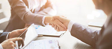 Zwei Personen schütteln sich über einem mit Dokumenten belegten Tisch die Hand, das Sonnenlicht scheint hell entgegen