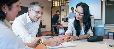 Drei Personen sitzen an einem Tisch, eine zeichnet etwas auf ein Blatt Papier