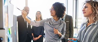 Frau in der Mitte erklärt etwas den umstehenden Personen an einer Pinnwand 